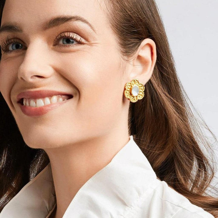 Gold flower-shaped clip-on earrings with a mother-of-pearl center, worn by a woman.