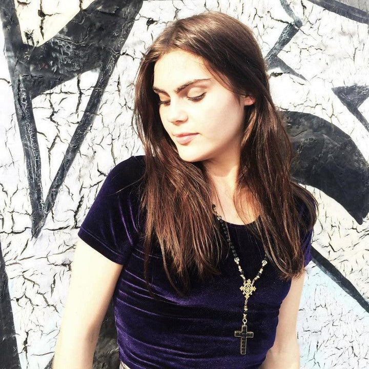 Woman wearing a dark velvet top and a long beaded necklace with a cross pendant against a graffitied wall.