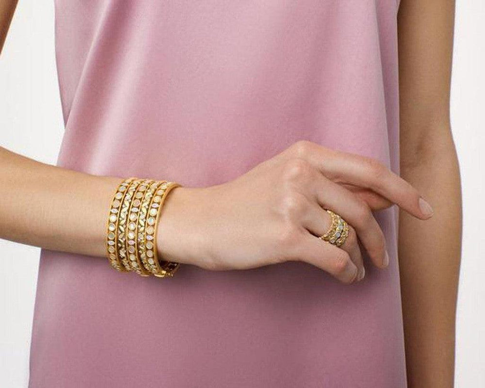 Gold bangles with iridescent stones, worn on a wrist against a dusty rose background.