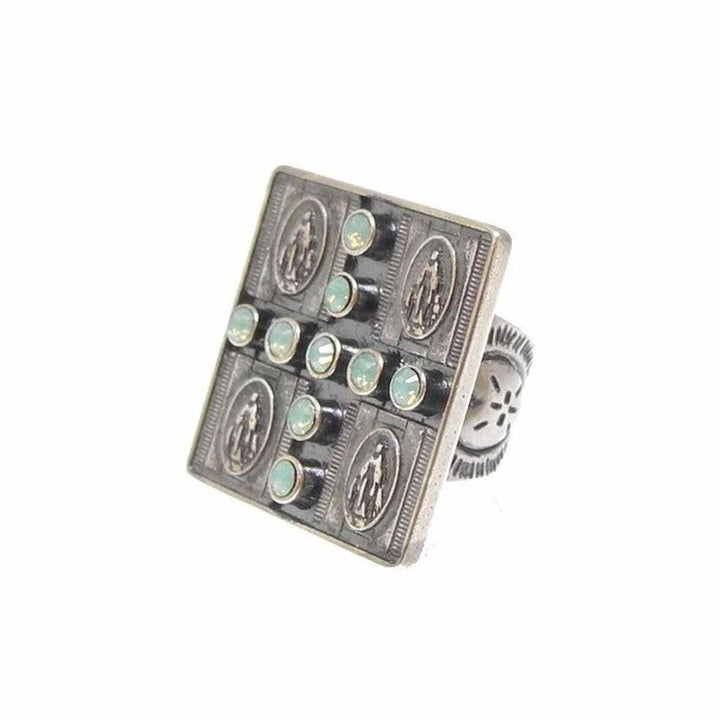 Square statement ring featuring a cross pattern of pale green stones, set against an antique silver-toned band with religious medallions.