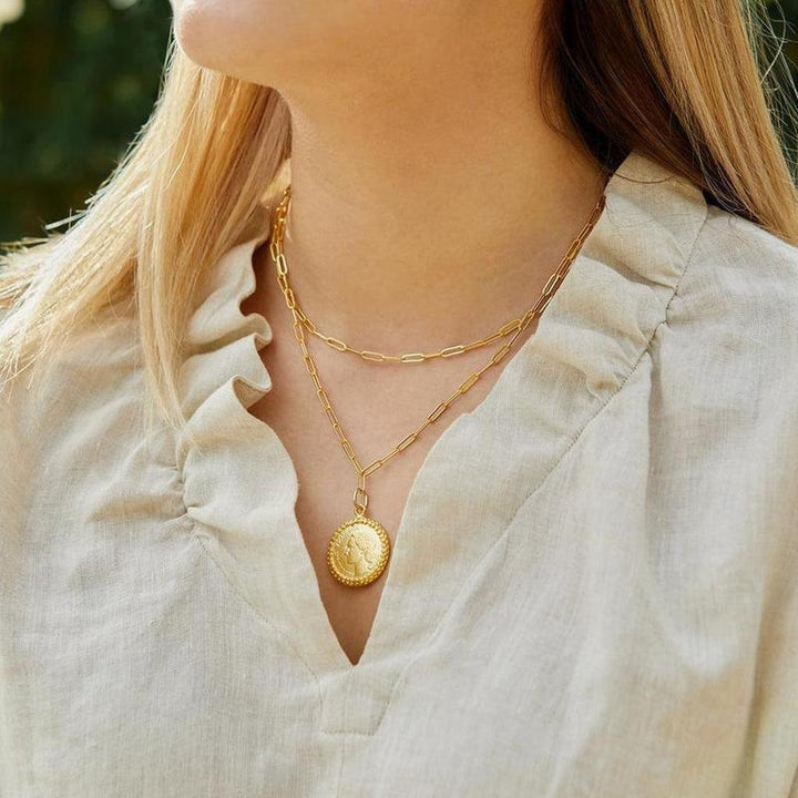 Gold coin pendant necklace with layered paperclip chain, styled on a beige linen top.
