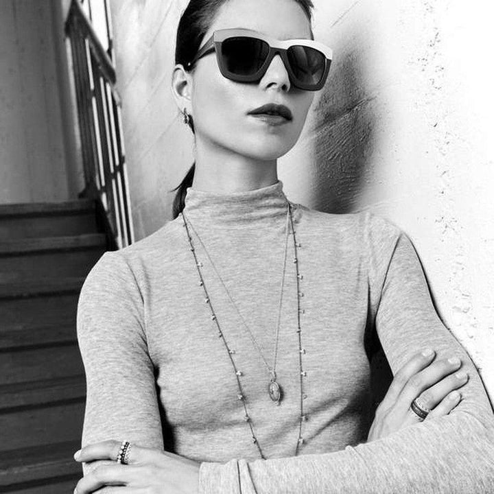 Model wearing layered necklaces and sunglasses, arms crossed, against a textured wall.