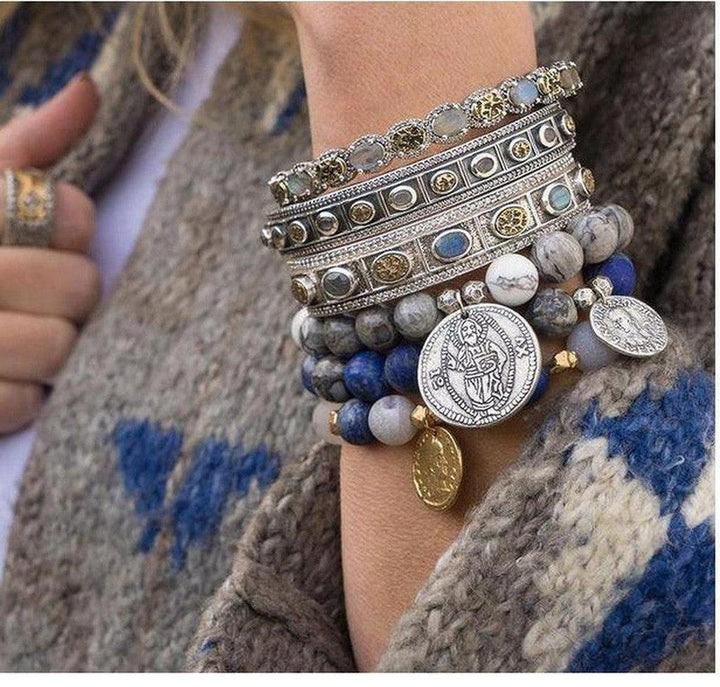 Silver hinged bangle bracelet layered with beaded bracelets; featuring gray, blue, and white stones.