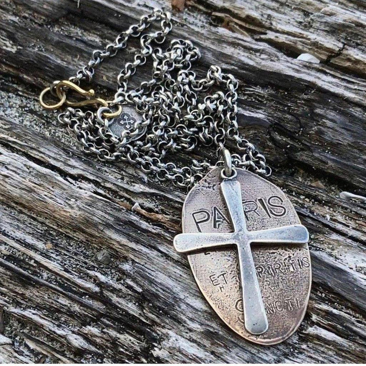 Silver chain necklace with an oval bronze pendant featuring a cross and engraved lettering, displayed on wood.
