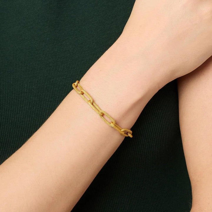 Gold paperclip chain bracelet on a wrist, shown against a dark green background.