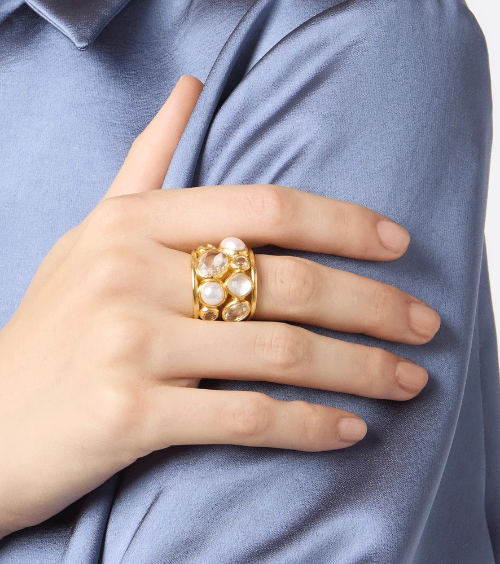 Gold statement ring featuring white pearls and gemstones; worn on a hand resting on a blue fabric.