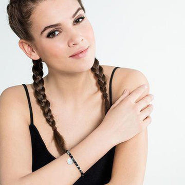 Woman with braided hair modeling a black beaded bracelet featuring a circular engravable charm.
