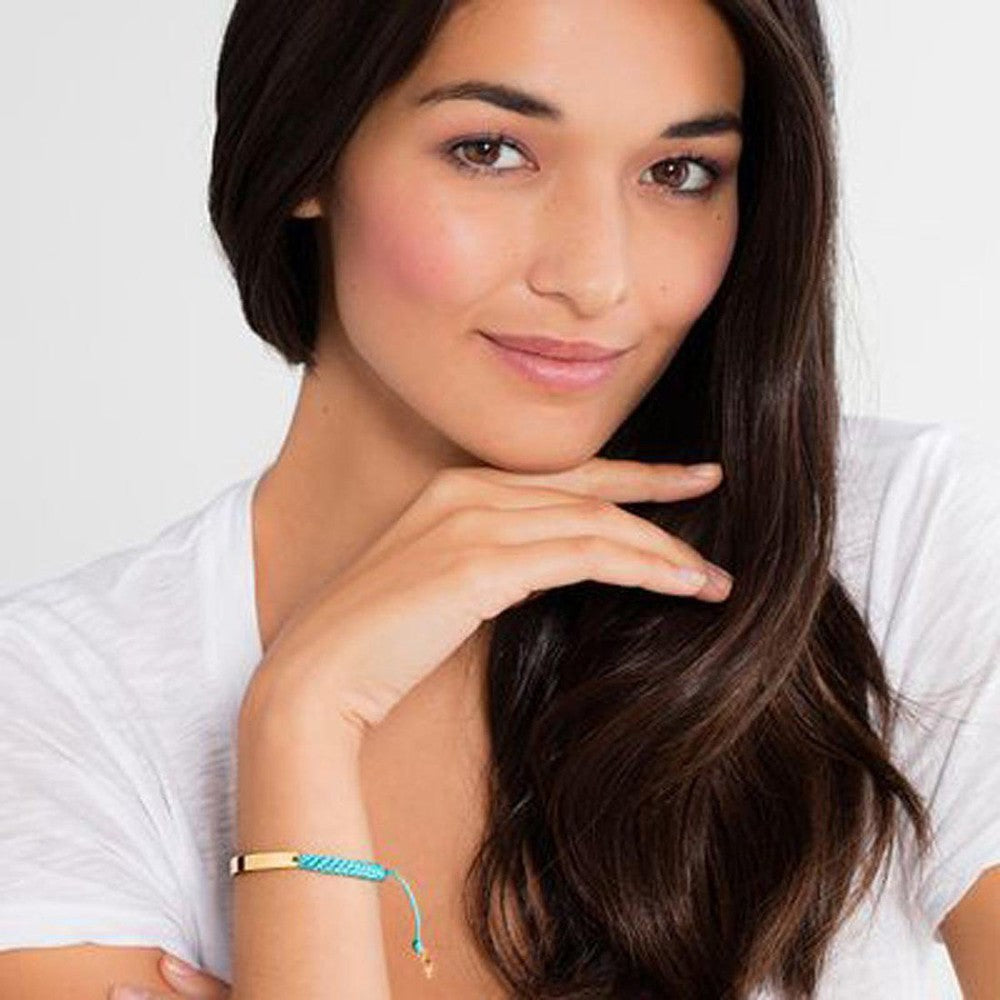 Gold bar bracelet with turquoise braided cord, worn by a woman.
