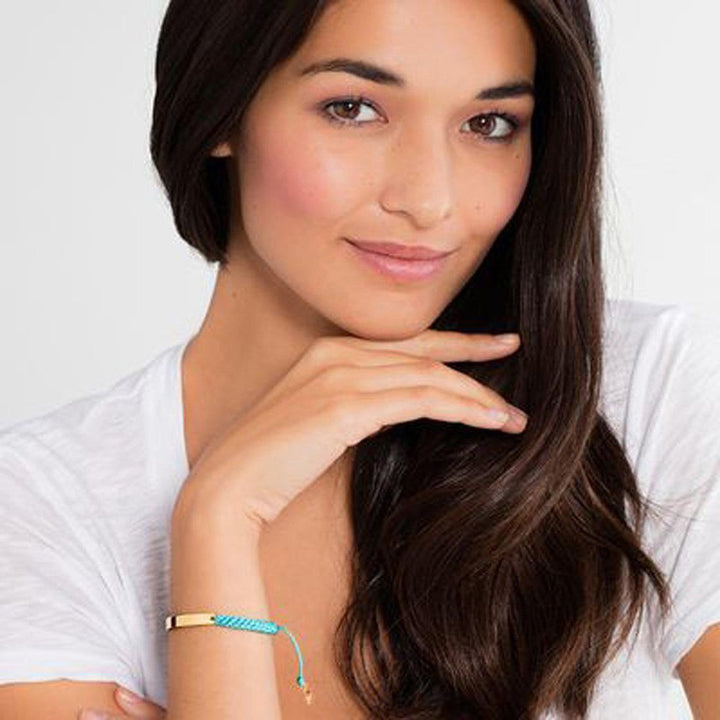 Gold bar bracelet with turquoise braided cord, worn by a woman.
