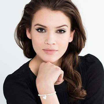 Woman's wrist wearing a delicate pearl bracelet with a small heart charm.
