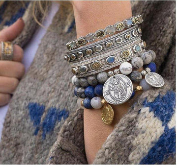 Silver hinged bangle bracelet layered with beaded bracelets; featuring gray, blue, and white stones.
