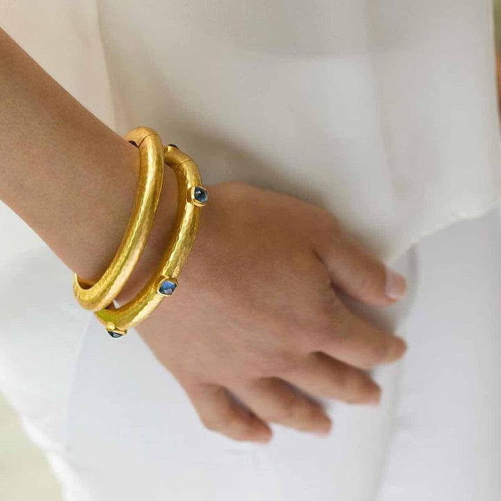 Gold hinged bangles adorned with blue gemstones, worn on a wrist.
